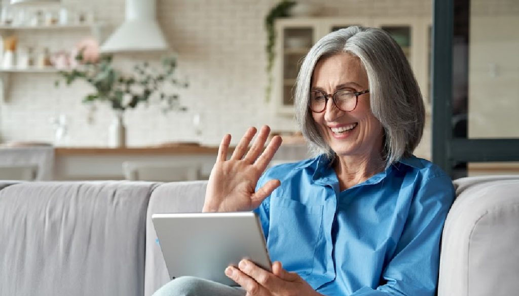 Happy 60s older mature middle aged adult woman waving hand holding digital tablet computer video conference calling by social distance virtual family online chat meeting sitting on couch at home. BillionGraves